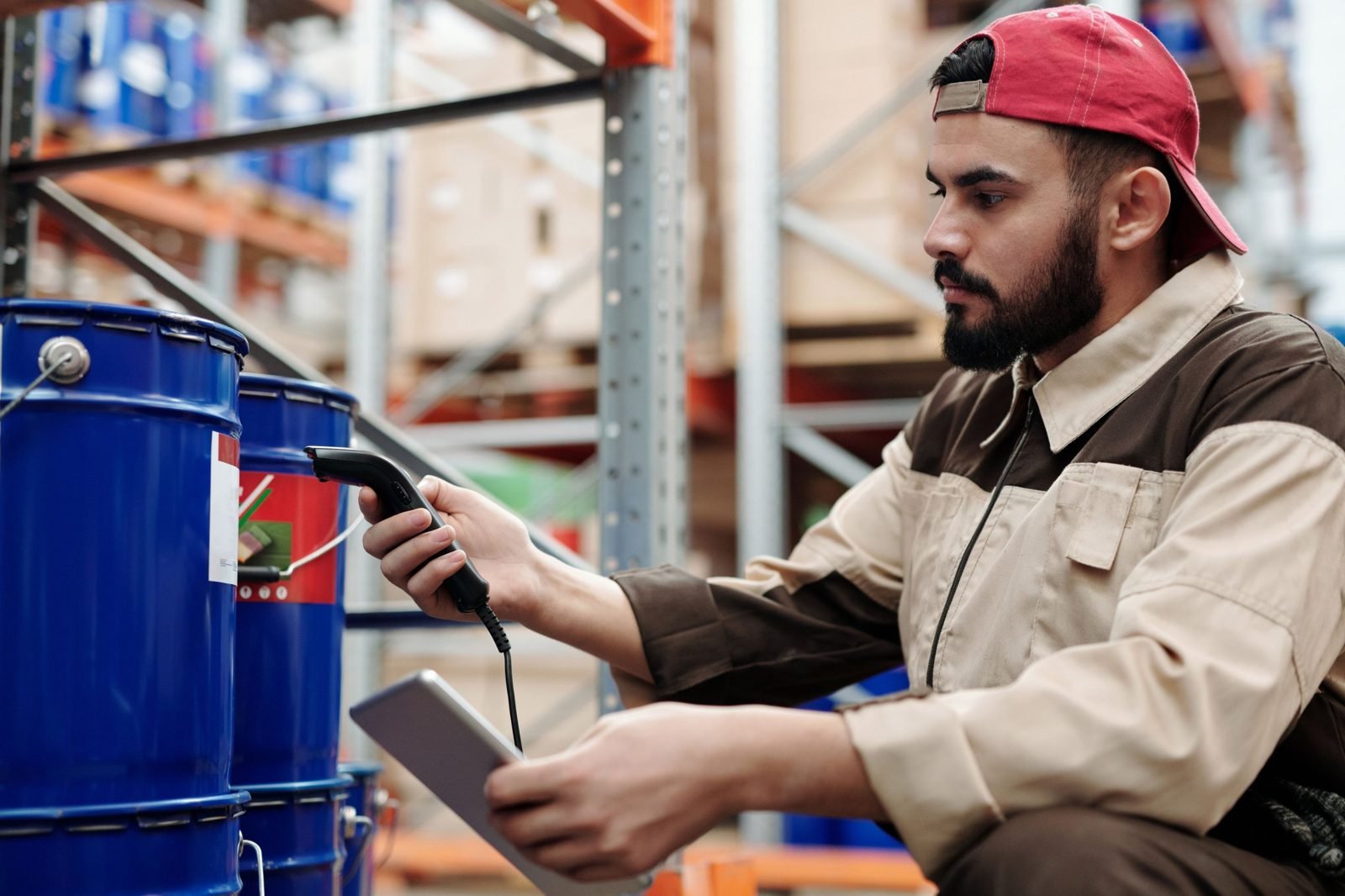 Industrial worker crouching to scan product labels on blue barrels with a handheld barcode scanner and tablet
