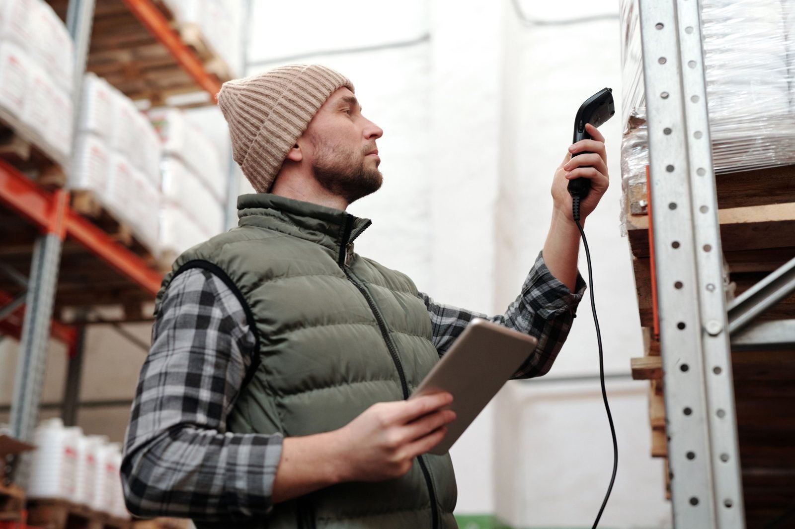 Warehouse employee holding a handheld barcode scanner toward palletized stock on metal racking while carrying a tablet